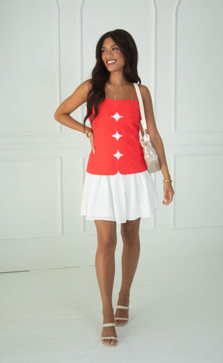 A woman modeling a sleeveless red mini dress with white details and a white skirt, paired with white heeled sandals and a white shoulder bag, standing against a white paneled wall.