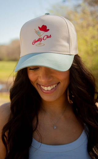 Woman wearing a beige and light blue trucker hat with a country club martini and cowboy hat graphic, outdoors.

