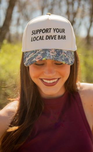 Woman wearing a camo trucker hat with 'Support Your Local Dive Bar' text outdoors.