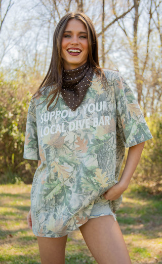 Woman wearing an oversized camouflage shirt with "support your local dive bar" outdoors