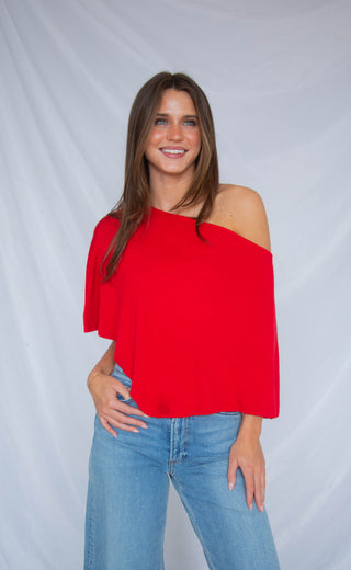 Woman wearing a red off-shoulder poncho top and blue jeans against a gray background