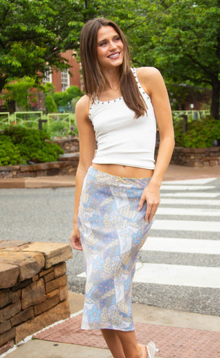 Woman wearing a white top and patterned skirt standing outdoors with greenery in the background