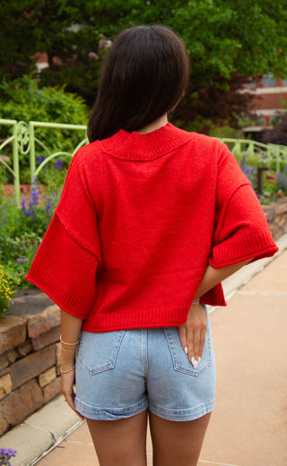 Person wearing a red sweater and denim shorts standing outdoors.