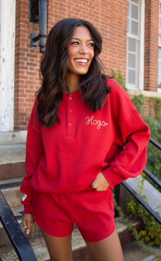 A woman smiling outdoors wearing a bright red long sleeve henley sweatshirt with white script embroidery reading 'Hogs' on the chest, paired with matching red shorts.