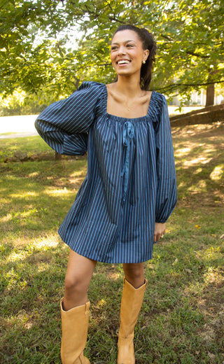 A woman wearing a blue and white vertically striped puff-sleeve mini dress with a tie neckline, paired with tan cowboy boots, standing outdoors in a grassy park with trees in the background.