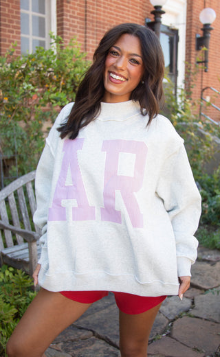 A woman smiling outdoors wearing an oversized light gray sweatshirt with large red and white striped 'AR' lettering on the front, paired with red shorts.