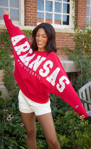 A woman posing outdoors in a bright red long-sleeve sweatshirt with 'ARKANSAS WOO PIG SOOIE' printed in white across the front, paired with white shorts.