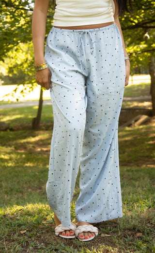 A person standing outdoors in a grassy park area wearing light blue wide-leg pajama pants with a small black polka dot pattern, a cream tank top, and white slippers.
