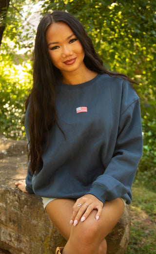 Woman wearing navy blue crewneck sweatshirt with embroidered American flag on the chest. 