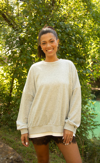 A woman wearing an oversized light beige crewneck sweatshirt and black lace shorts, standing outdoors in front of a lush green background.