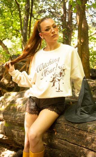 A woman wearing a cream sweatshirt with a 'Witching Hour!' graphic sits on a log in a forest. She is wearing a black witch hat and yellow boots.