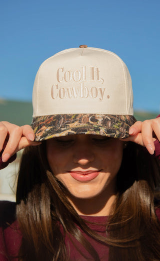 Person wearing a camo trucker hat with 'Cool It, Cowboy.' text against a blue sky