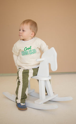 Child sitting on a white rocking horse against a beige background wearing a graphics sweatshirt that reads "cool kids club" 