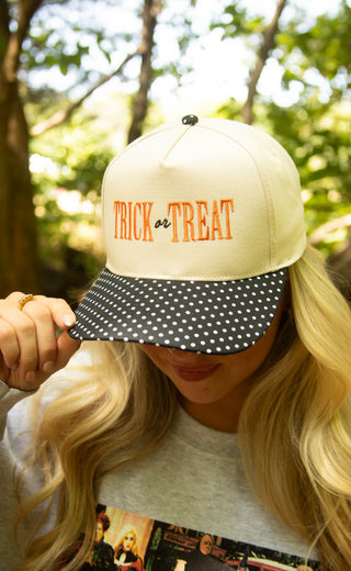 A woman outdoors wearing a cream baseball cap with orange embroidered text reading 'TRICK or TREAT' and a black brim covered in white polka dots.