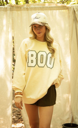 A woman wearing a beige oversized sweatshirt with 'BOO' printed in varsity block letters on the front, paired with dark plaid shorts and a camouflage baseball cap, standing in front of a draped cream fabric backdrop.