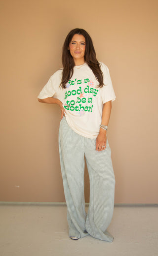 Woman wearing a cream graphic t-shirt with green text that reads "it's a good day to be a mother" and light gray checkered pants against a beige wall.