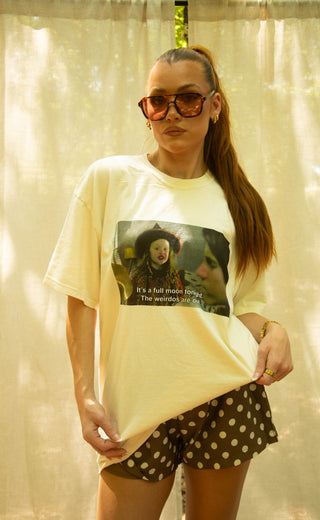 A woman wearing a cream-colored graphic t-shirt with a meme print and brown polka-dot shorts, standing in front of a beige curtain backdrop.