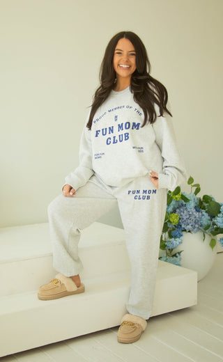 Woman wearing a 'Fun Mom Club' sweatshirt and matching sweatpants sitting on a couch with flowers in the background.