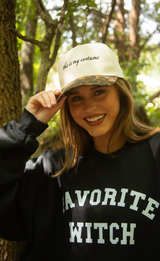 A woman outdoors in a wooded setting wearing a black sweatshirt with 'FAVORITE WITCH' printed in white and a white baseball cap with 'this is my costume' written on it.