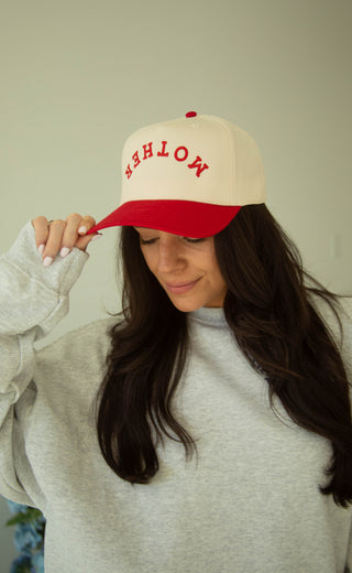 Woman wearing a beige and red trucker hat with 'MOTHER' text on a plain background