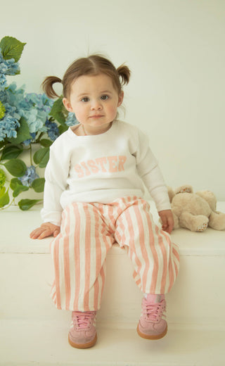 Child wearing a white "sister" sweatshirt and pink striped pants sitting on a white surface with flowers and a teddy bear in the background.