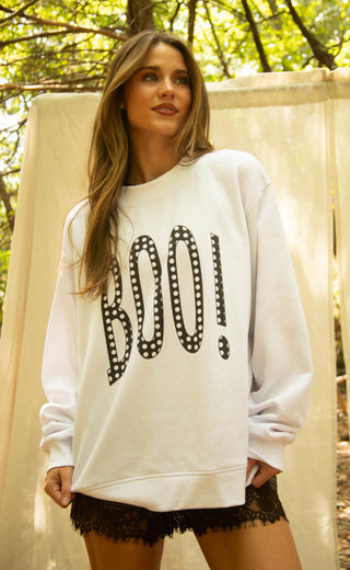 A woman wearing an oversized white crewneck sweatshirt with 'BOO!' printed across the front in a black dotted font, posed outdoors in front of a draped fabric backdrop.