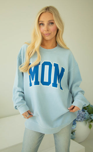 Woman wearing a light blue sweatshirt with 'MOM' in blue letters, standing against a white background.