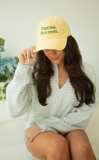Woman wearing a yellow cap with 'Trust me, I'm a mom' text, sitting on a white surface.