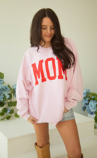 Woman wearing a pink sweatshirt with 'MOM' in red letters, standing indoors with flowers in the background.