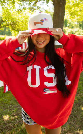 Person wearing a red 'USA' sweatshirt and white cap with a dachshund design outdoors.