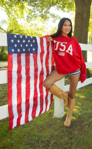 Person wearing a red 'USA' sweatshirt holding an American flag outdoors.