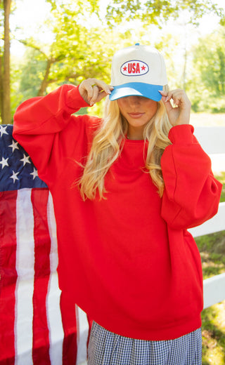 Person wearing a red sweatshirt and blue and white cap with 'USA' text, standing in front of an American flag.