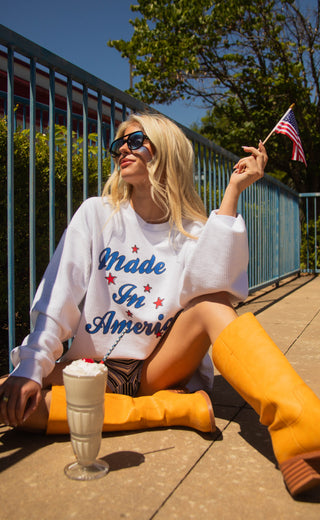 Woman sitting outdoors holding a small American flag, wearing a 'Made in America' sweatshirt.