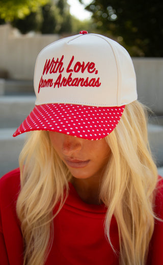 A blonde woman wearing a white and red trucker-style cap with red embroidered text reading 'With Love, from Arkansas' and a red brim covered in white polka dots, photographed outdoors with a blurred background.