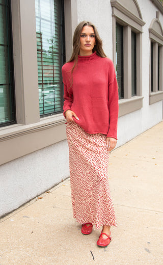 A woman stands on a sidewalk in front of a building, wearing a coral pink knit mock-neck sweater and a matching pink and white patterned maxi skirt with red shoes.