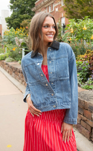 A woman wearing a cropped light-wash blue denim jacket with a black collar, styled over a red top and a red and white striped skirt, standing outdoors in front of a brick building and garden.