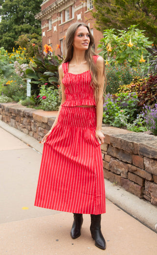 A woman modeling a red two-piece set with thin white vertical stripes, featuring a sleeveless crop top and a matching maxi skirt, paired with black ankle boots, set against a garden and brick building background.
