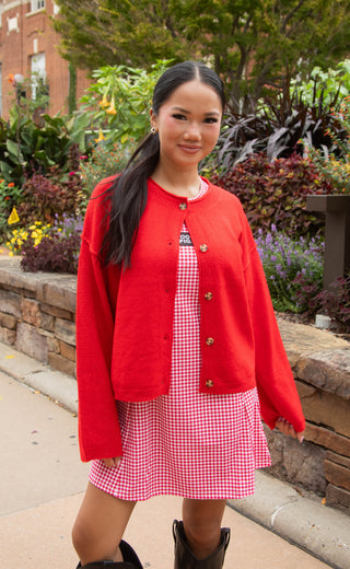 A woman wearing a bright red knit cardigan over a red and white gingham dress, standing outdoors in front of a stone wall and green plants.