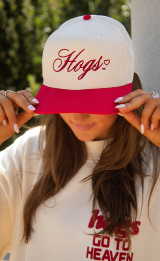A woman wearing a white and bright pink baseball cap with 'Hogs' embroidered in pink script on the front, holding the brim down to her eyes. She is also wearing a matching white t-shirt with 'hogs GO TO HEAVEN' printed in red.