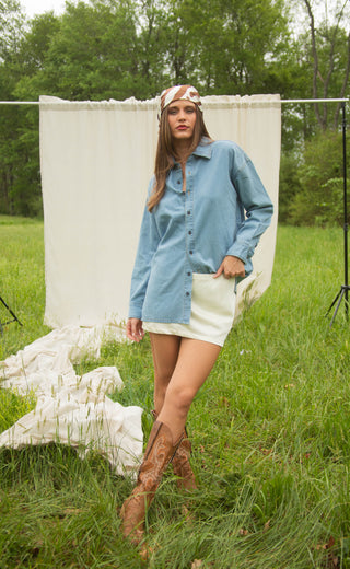 Woman in a blue shirt and white skirt standing in a grassy field with a white backdrop.