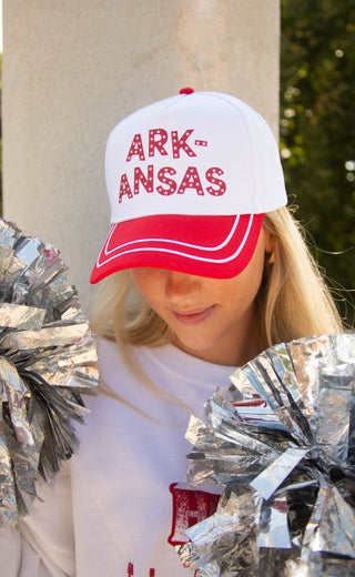 A woman wearing a white and red baseball cap with 'ARKANSAS' in red letters, holding silver foil pom-poms in front of her. The background shows a beige stone pillar and green foliage.