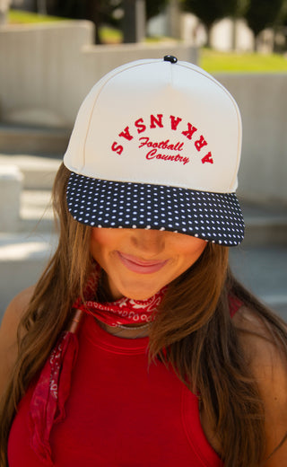 A woman wearing a white baseball cap with red embroidered text reading 'Arkansas Football Country' and a navy polka-dot visor, photographed outdoors with a blurred background.