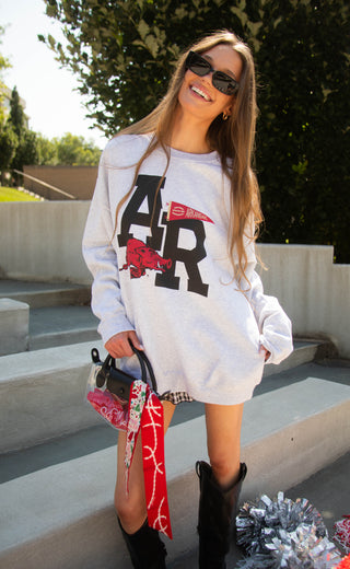 A woman wearing a white long-sleeve graphic sweatshirt with a large black 'AR' and red hog logo, styled with black boots, sunglasses, and a red patterned handbag, standing outdoors on concrete steps.