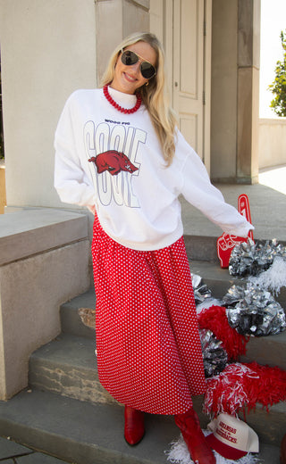 A woman stands on outdoor stone steps wearing a white sweatshirt with a large SOOIE graphic and a red polka-dot skirt, accessorized with red boots, a red beaded necklace, and sunglasses. 