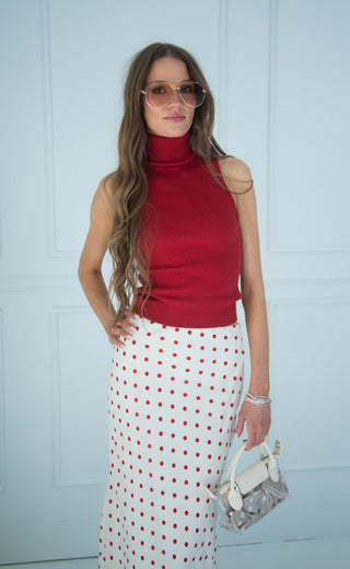 A woman modeling a sleeveless red turtle-neck top and a white maxi skirt with red polka dots, holding a small white handbag against a light blue paneled wall.