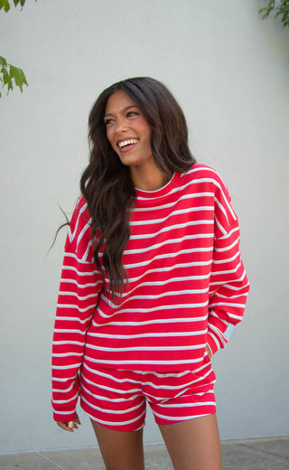 A woman smiling and looking to the side while wearing a red and blue horizontally striped long-sleeve top and matching shorts against a light gray wall.