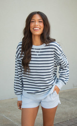 A woman smiling and wearing a navy and white horizontally striped long-sleeve crewneck sweatshirt with light blue shorts, standing against a neutral beige wall.