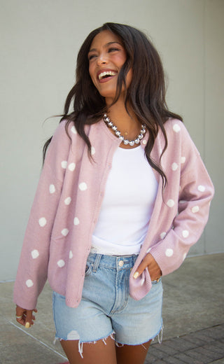 A woman wearing a pink cardigan with white polka dots over a white top and denim shorts, smiling and looking to the side in an outdoor setting.