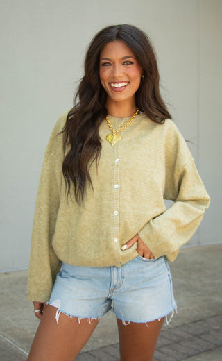 A woman smiling and wearing a fuzzy, oversized olive button-up cardigan with light-wash denim shorts, standing against a neutral gray wall.