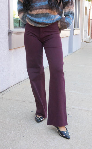 A person wearing a pair of high-waisted, wide-leg flared pants in a deep burgundy or plum purple color, paired with a striped knit sweater and black polka-dot pointed flats, standing on a concrete sidewalk.